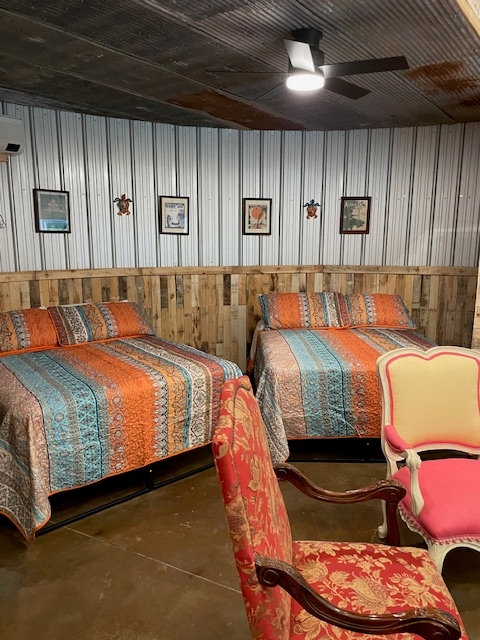 Eclectic bedroom area within a grain bin featuring two queen beds with vibrant orange and blue patterned linens, set against corrugated metal and wood-paneled walls with framed farm-themed art.