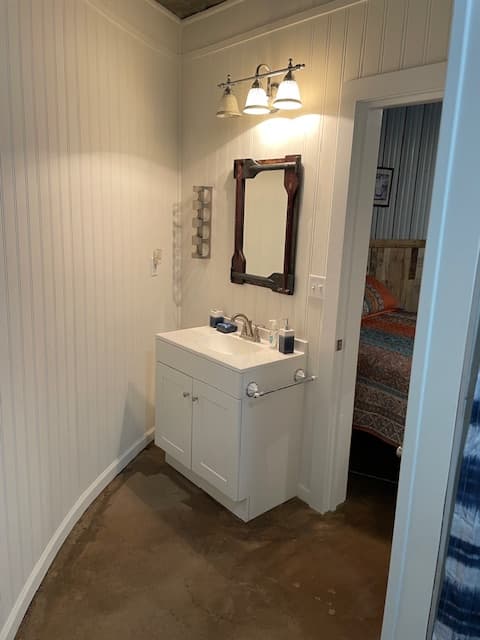 A white single-sink vanity with a dark-framed mirror and three-bulb vanity light, set against light-colored vertical paneled walls in a bathroom with a concrete floor.