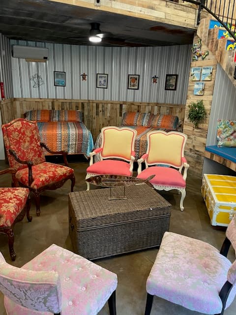 An eclectic seating area inside a grain bin, featuring vintage floral and pink velvet chairs arranged around a wicker trunk, with beds and corrugated metal walls in the background.