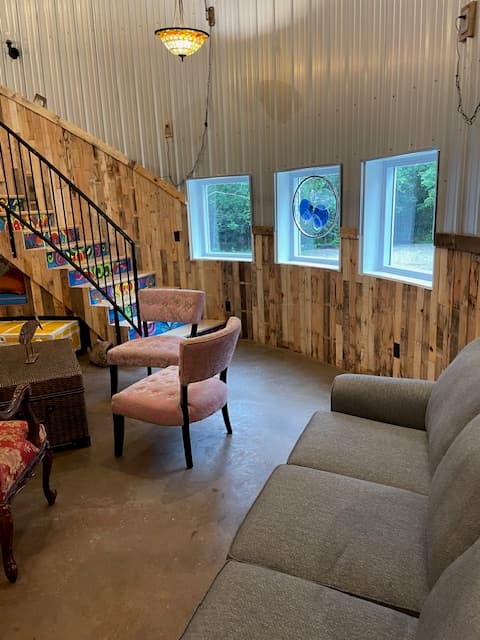 A circular living area inside a grain bin, featuring three small square windows, a grey sofa, and pink tufted chairs. The space is finished with wood-plank wainscoting and corrugated metal walls.