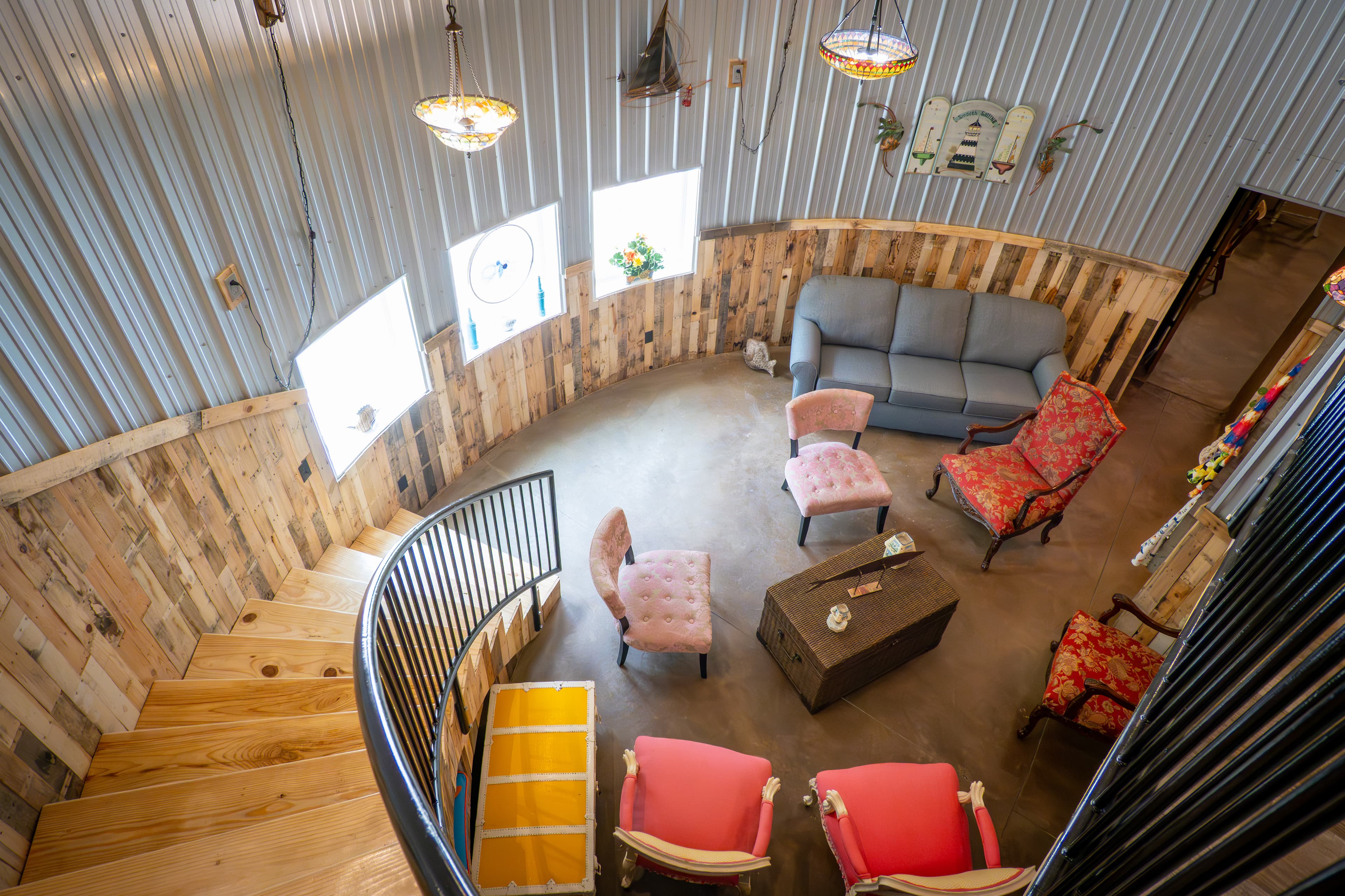High-angle view of a circular living area inside a converted grain bin, featuring corrugated metal walls, wood paneling, a spiral staircase, and eclectic seating around a central coffee table.