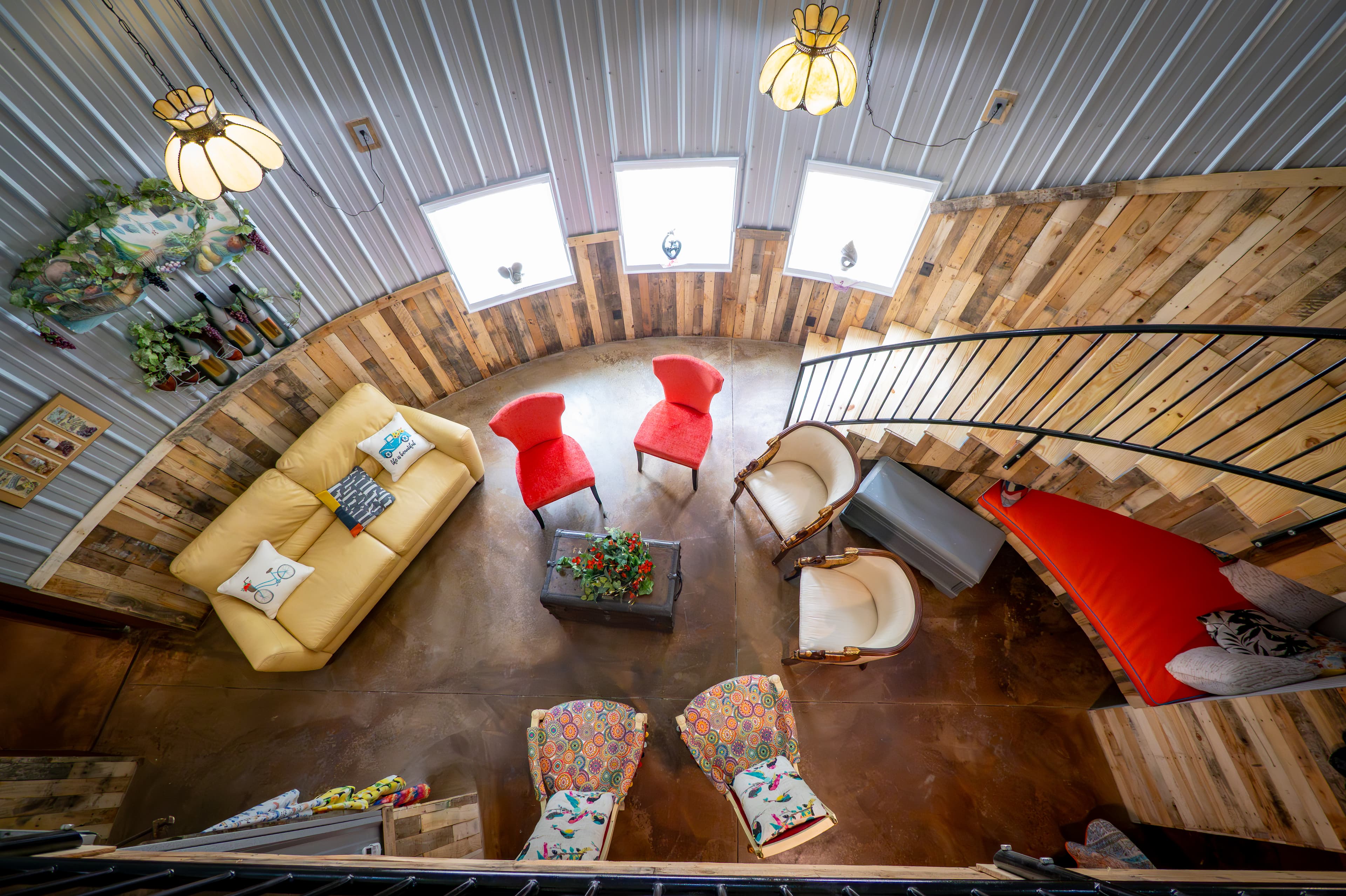 High-angle view from a loft looking down into a circular living area with corrugated metal walls and wood wainscoting. The space features an eclectic mix of seating, including a yellow sofa, two bright red chairs, and vintage patterned armchairs arranged around a central trunk. A wooden staircase with a black metal railing and a red built-in daybed underneath follows the curve of the wall.