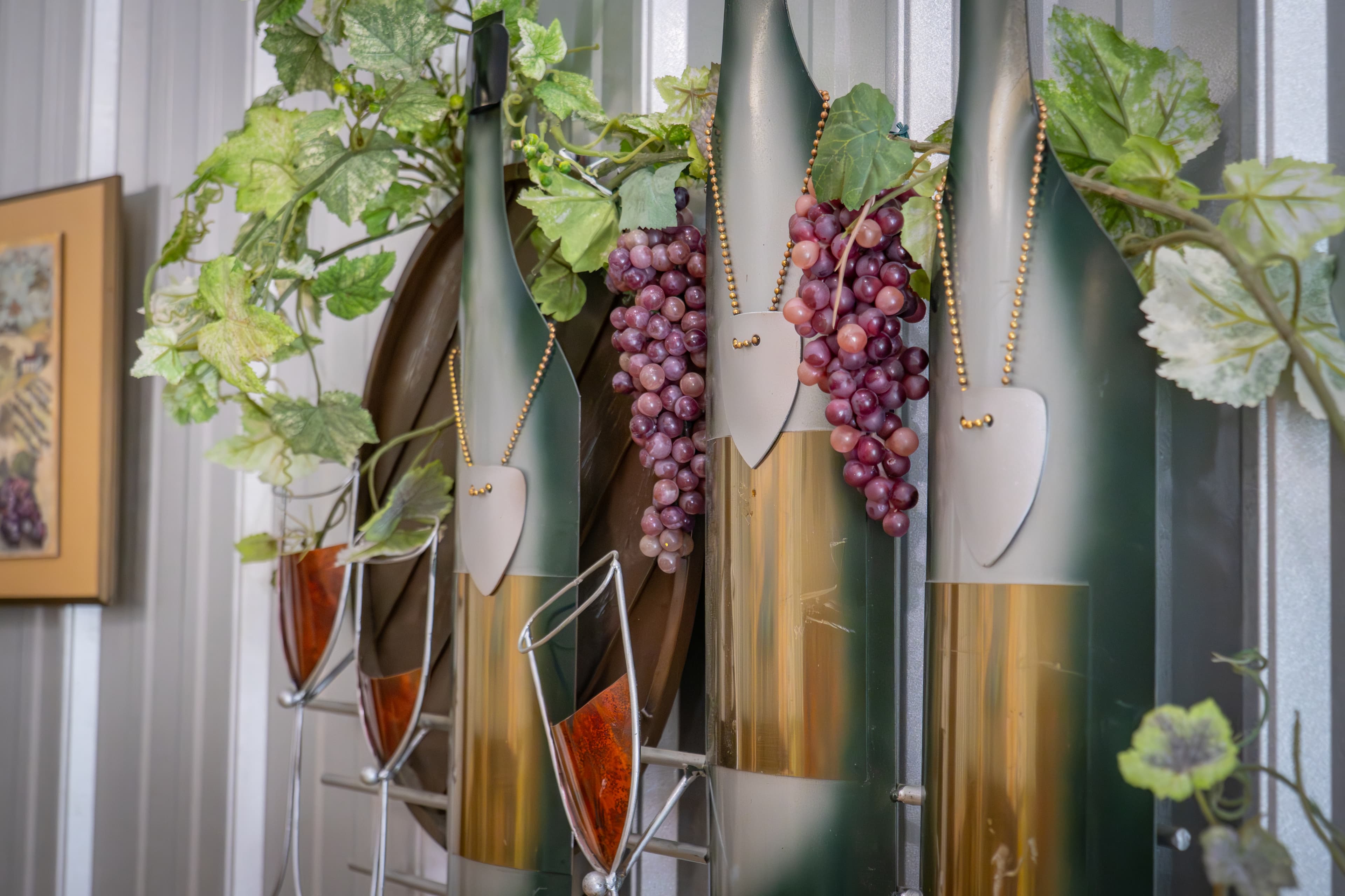 Wall-mounted wine-themed art featuring green and gold stylized metal bottles adorned with artificial grape clusters and vine leaves, set against a corrugated metal wall.