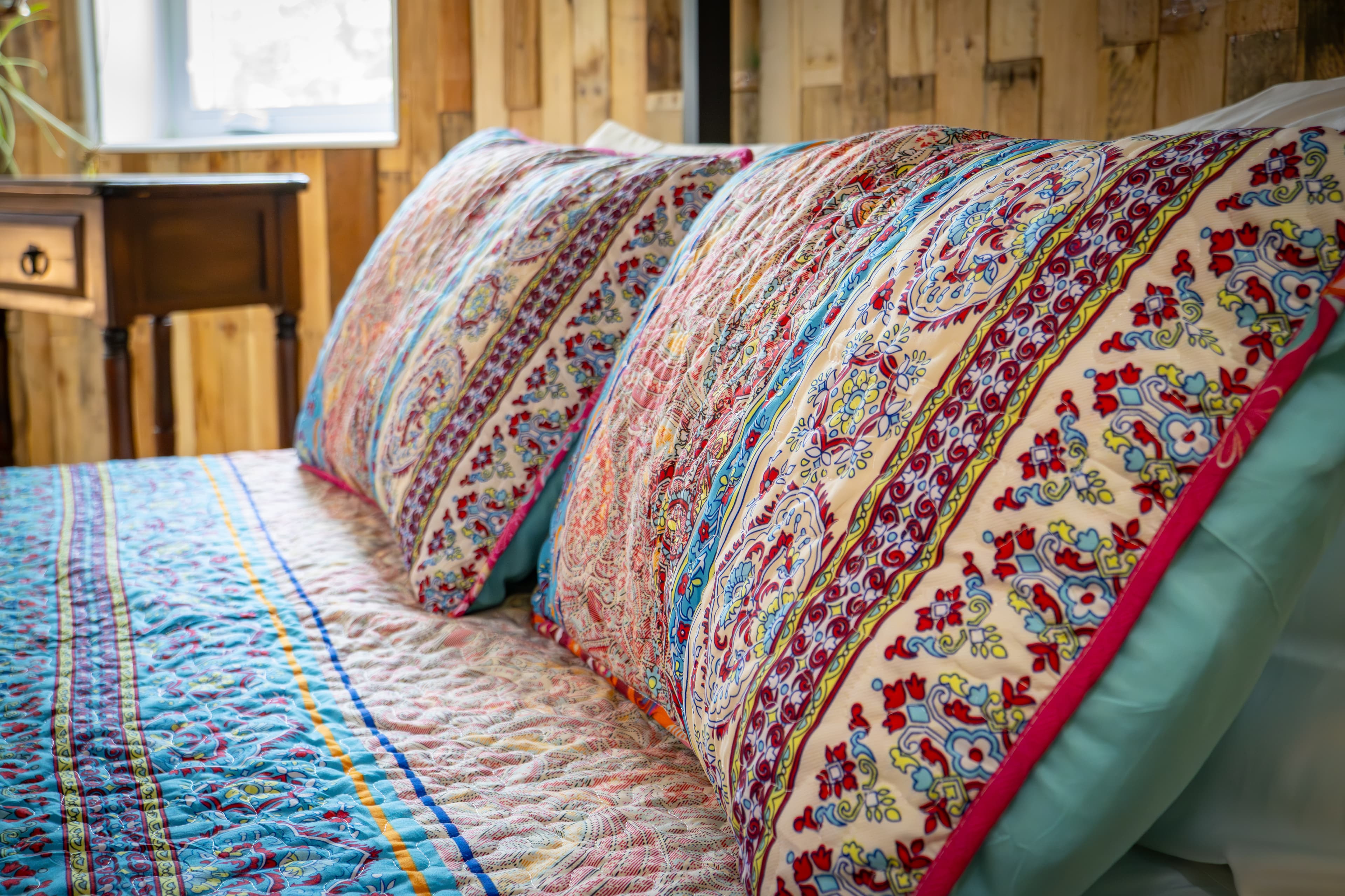 Close-up of decorative pillows and bedding featuring an intricate, multi-colored Bohemian pattern with vibrant pink, teal, and gold accents, set in a room with rustic wood-paneled walls.