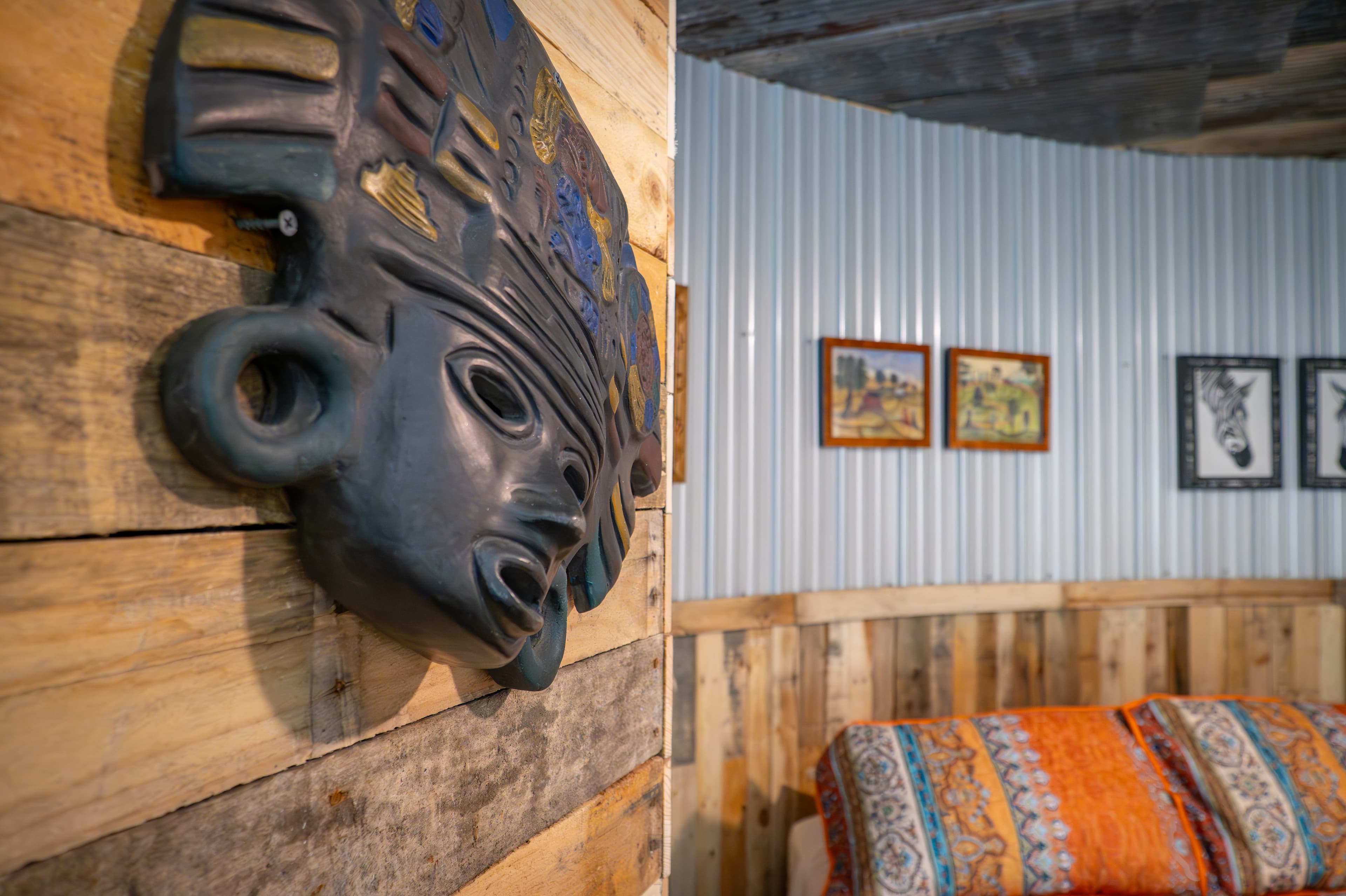 A decorative, tribal-style wall mask in dark blue with gold and blue accents is mounted on a rustic wood-plank wall. In the blurred background, two queen beds with vibrant orange and blue patterned linens are visible against a corrugated metal wall adorned with small framed pictures.