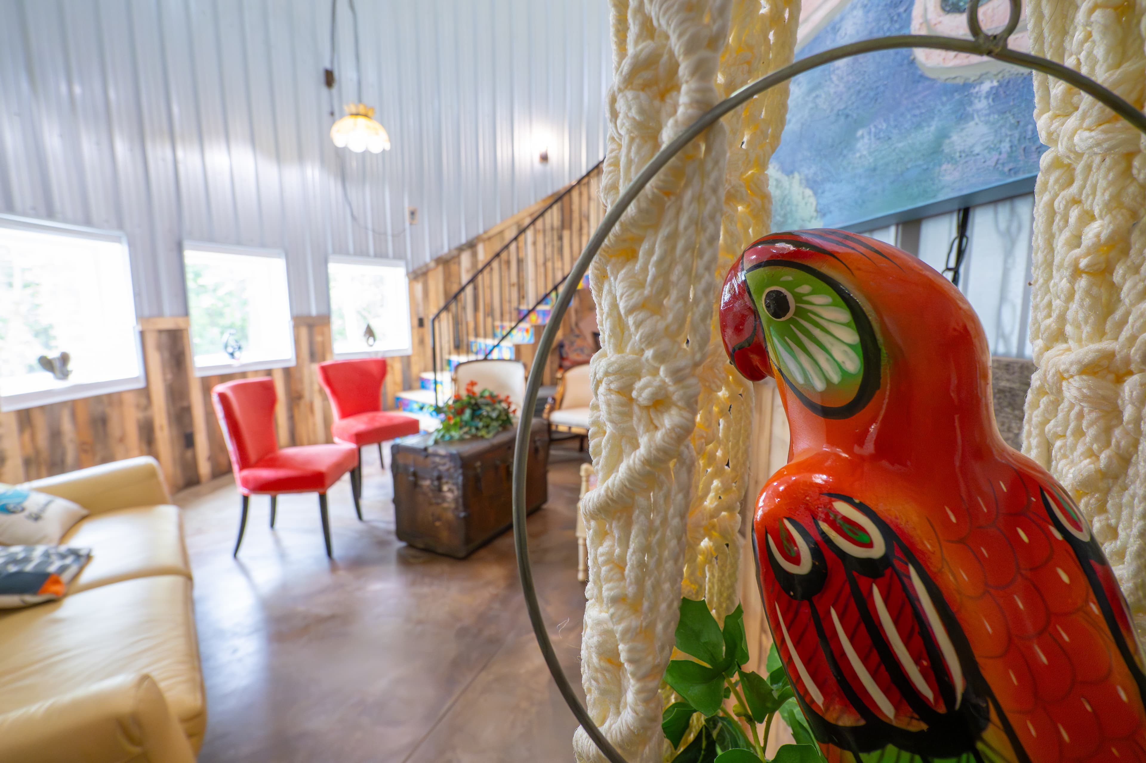 A colorful, hand-painted wooden parrot figurine sits in the foreground, with a blurred view of a vibrant grain bin living area in the background. The background reveals red chairs, a tan sofa, and a rustic trunk coffee table against corrugated metal and wood-plank walls.