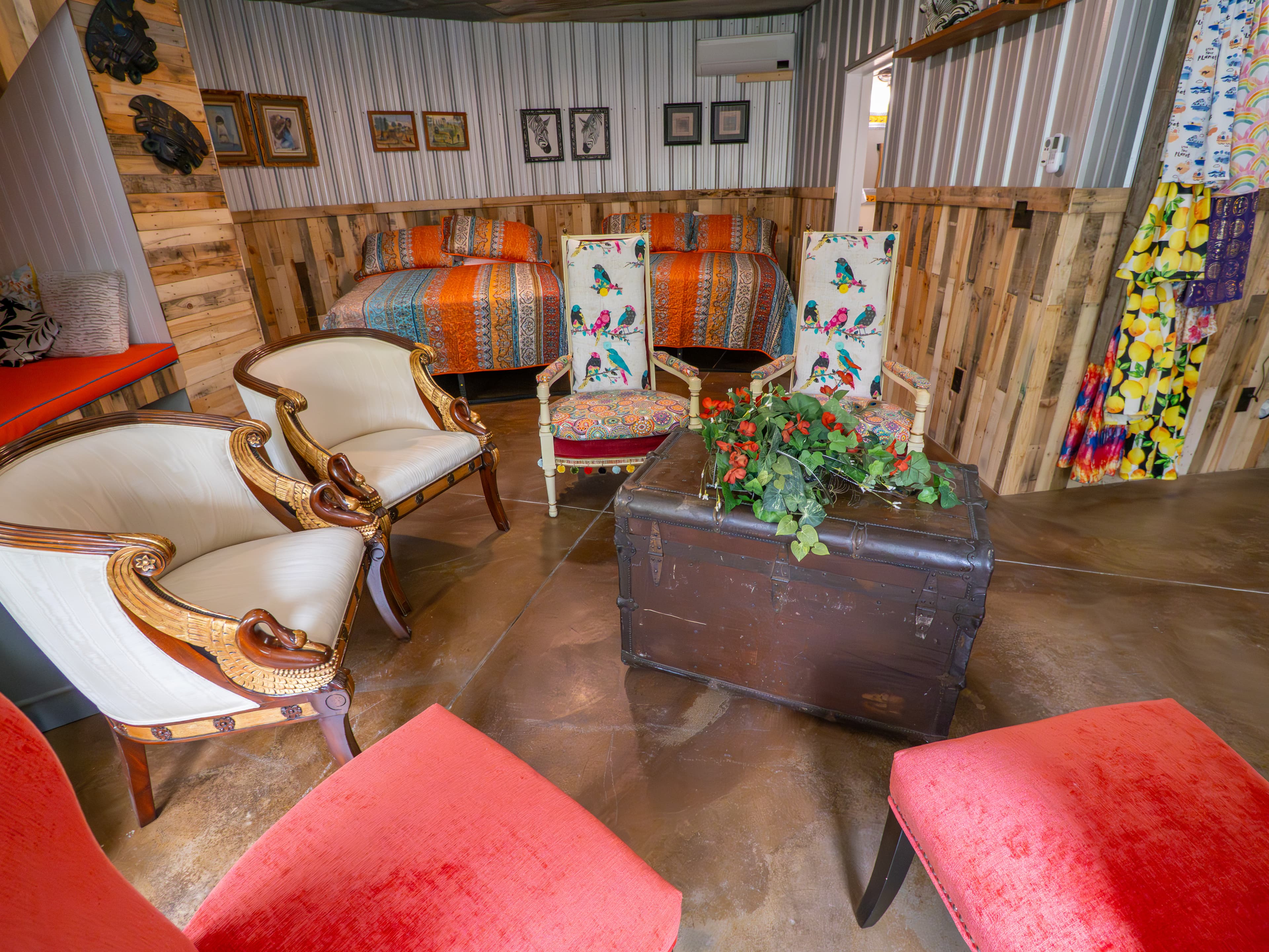 Eclectic seating area on the lower level of a circular grain bin, featuring vintage armchairs with bird-patterned upholstery and red velvet stools arranged around a rustic wooden trunk. In the background, two queen beds with colorful striped linens are nestled against wood-paneled and corrugated metal walls.