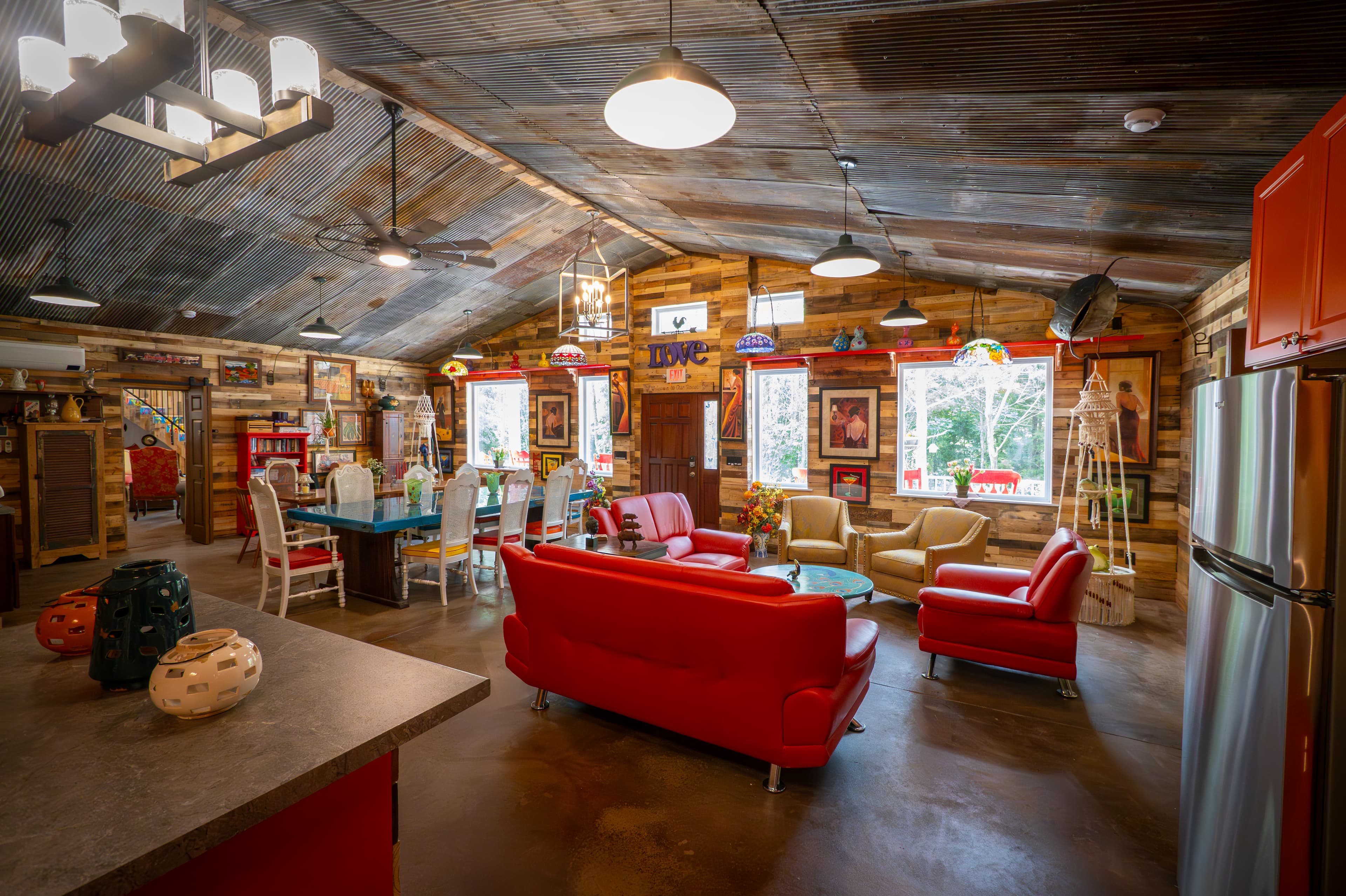 A wide-angle view of "The Roost" common area, featuring a spacious kitchen with bright orange cabinets and stainless steel appliances. The living space is filled with vibrant red leather sofas and armchairs, a turquoise peacock-themed coffee table, and large windows that fill the rustic, wood-paneled room with natural light.