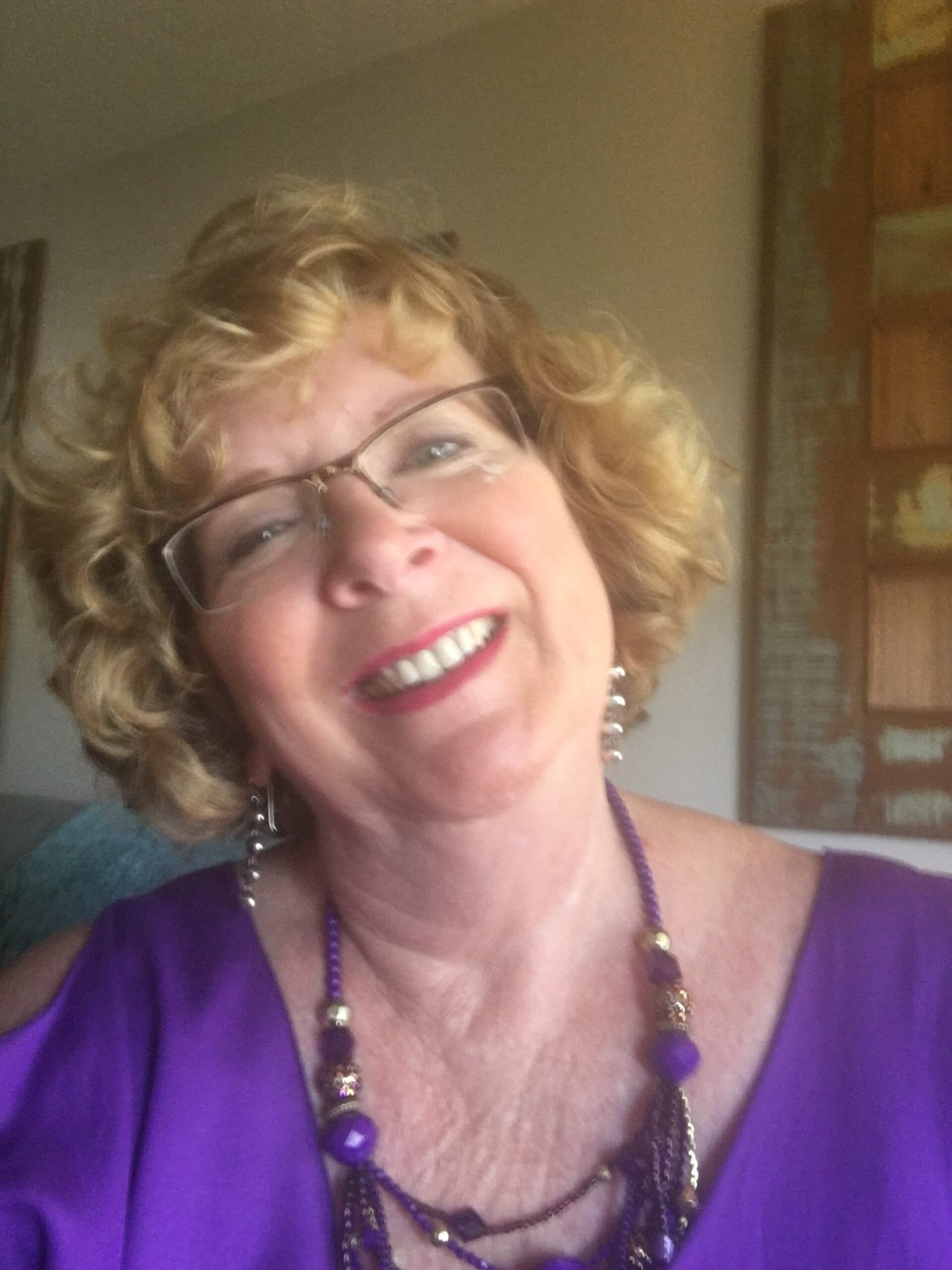 A smiling woman with curly hair wearing a purple top and necklace poses for a selfie.