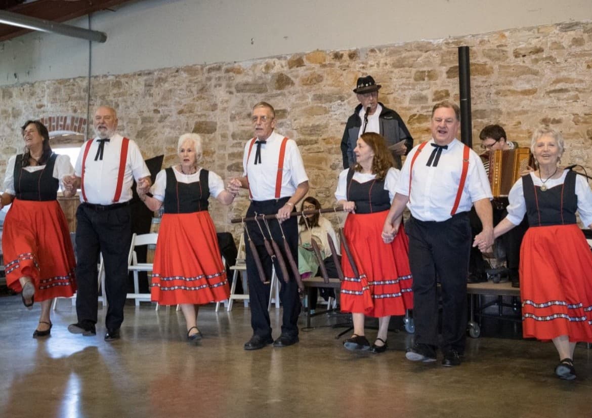 A group of dancers in traditional attire perform together, holding hands in a lively setting.