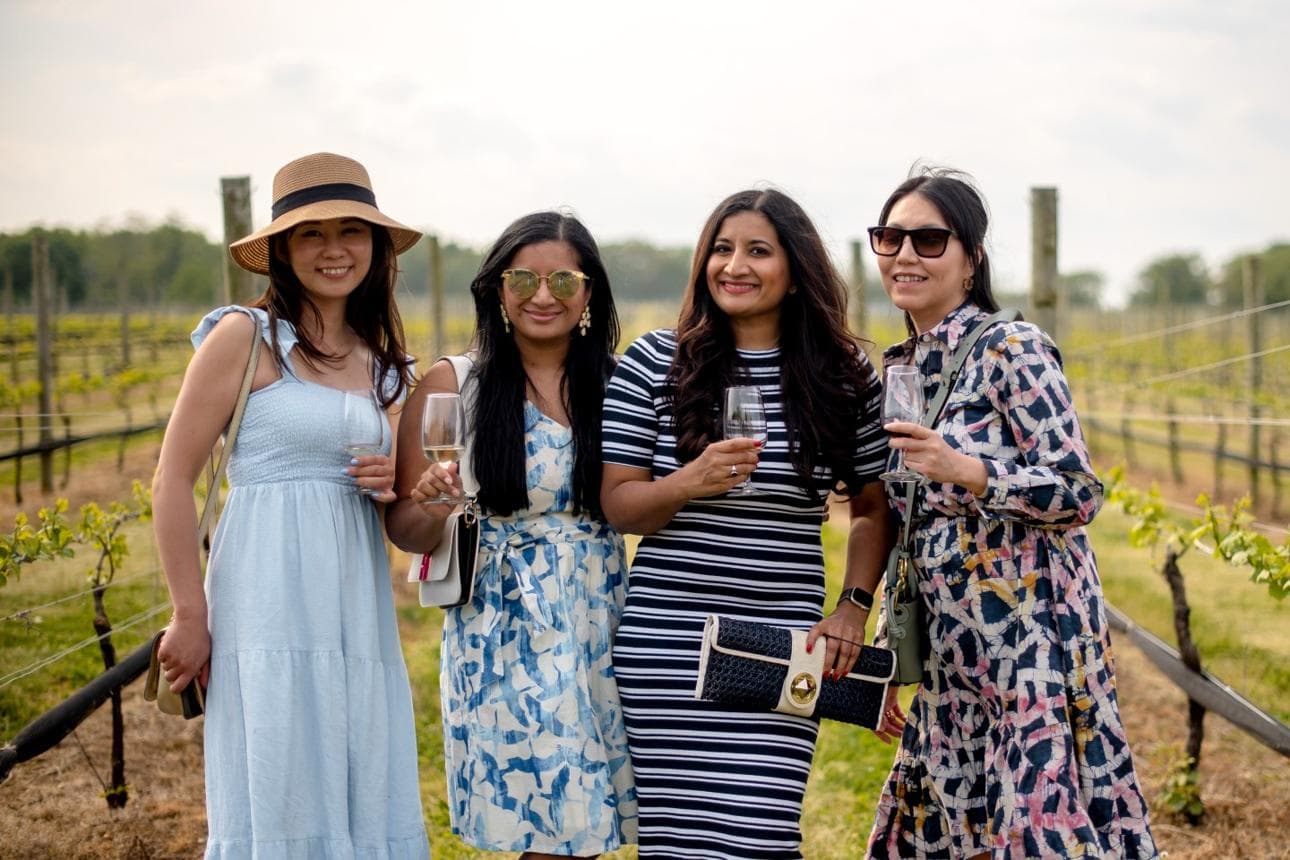 Four women in stylish outfits pose with glasses of wine in a vineyard.