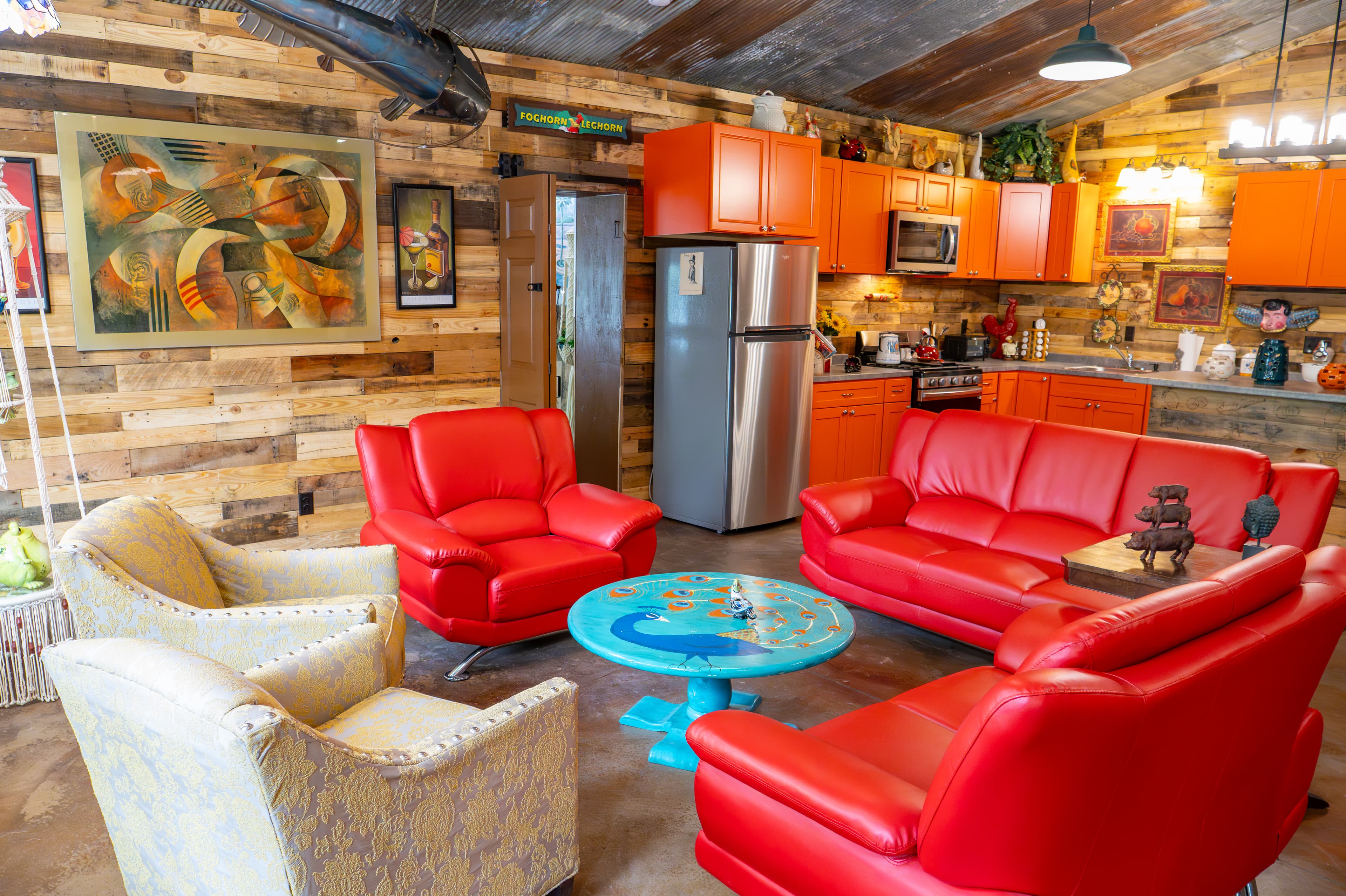 A vibrant, eclectic common area in the Roost featuring bright red leather sofas and armchairs, a turquoise peacock-themed coffee table, and a kitchen with bold orange cabinetry. The room is decorated with abstract art and rustic wood-paneled walls.