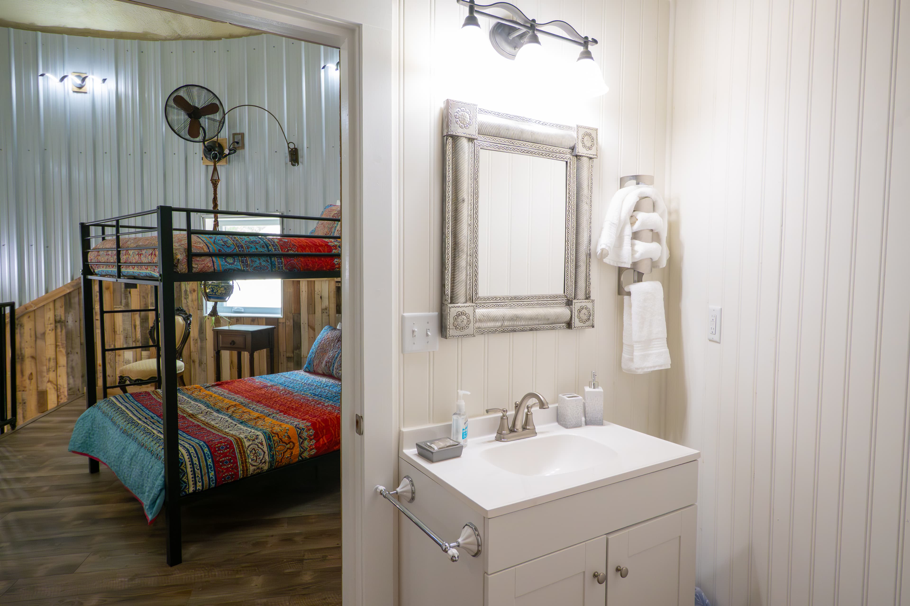 A white single-sink vanity with a distressed silver-framed mirror and a three-bulb light fixture, viewed from an open doorway that reveals a black metal bunk bed with colorful striped bedding in the adjacent room.