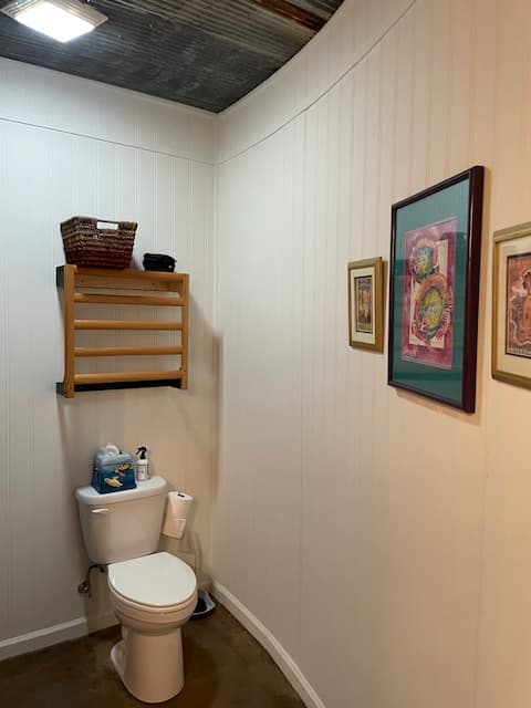 A white toilet in a bathroom with light-colored, vertically paneled walls and a concrete floor. Above the toilet is a wooden towel rack with a basket on top, and framed art hangs on the adjacent wall.