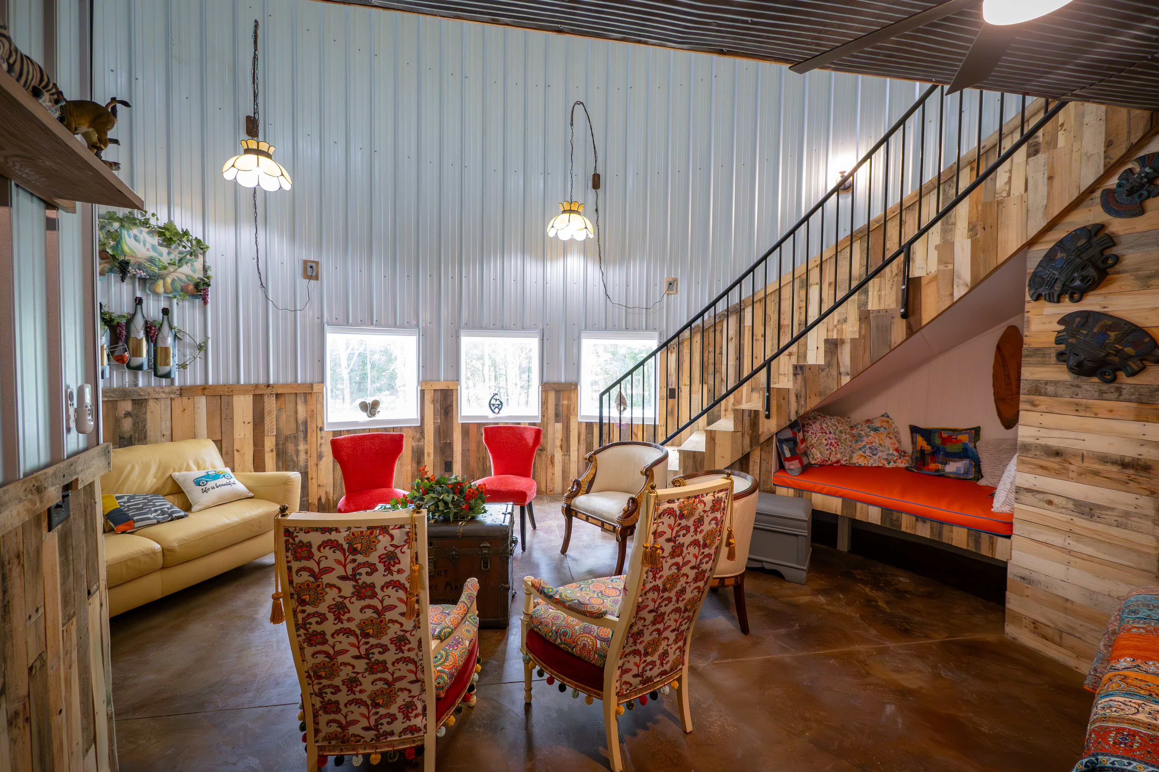 A vibrant, eclectic living space inside a circular grain bin featuring high corrugated metal walls and wood-plank wainscoting. The room is filled with colorful vintage armchairs, a tan sofa, and a built-in orange daybed tucked under a wooden staircase, all arranged around a central rustic trunk coffee table.