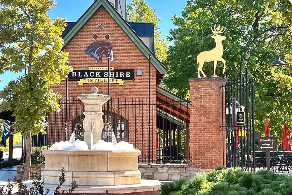 A brick distillery building with a golden deer statue and a fountain in front, surrounded by trees.