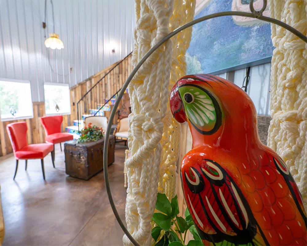 A colorful parrot statue hangs in a cozy interior with red chairs and wooden accents.
