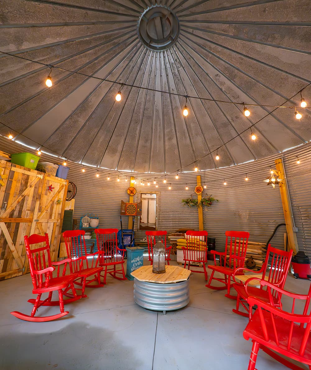 A cozy indoor space featuring red rocking chairs arranged in a circle around a wooden table, with string lights illuminating the metal dome ceiling.