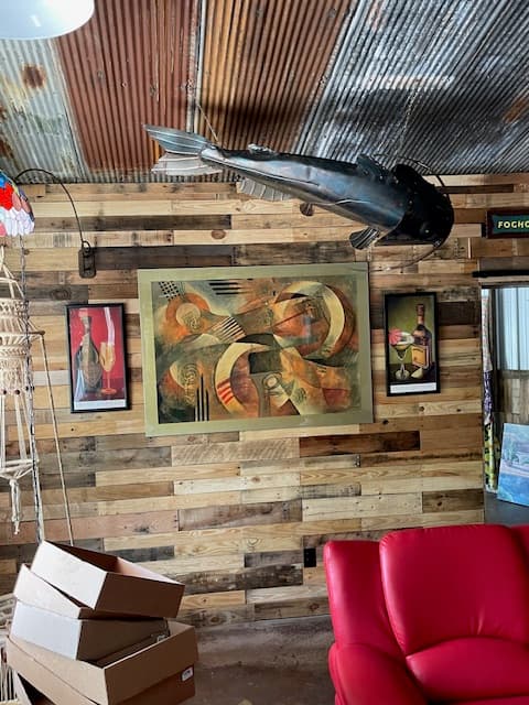A rustic wall featuring abstract art, framed pictures, and a suspended fish sculpture, alongside a red chair and storage boxes.