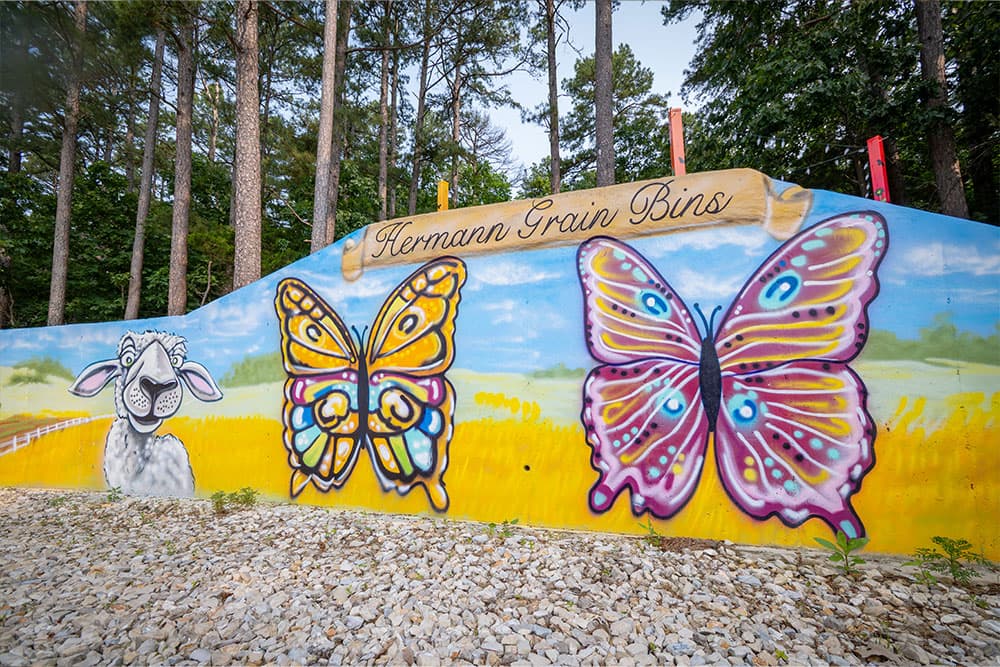 Colorful mural featuring a sheep and butterflies with the text "Hermann Grain Bins" above.