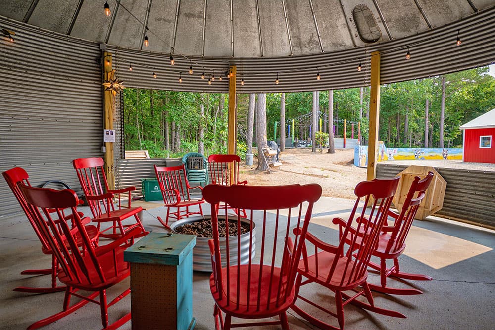 A circle of red rocking chairs surrounds a fire pit in a rustic, open shelter with forest views.