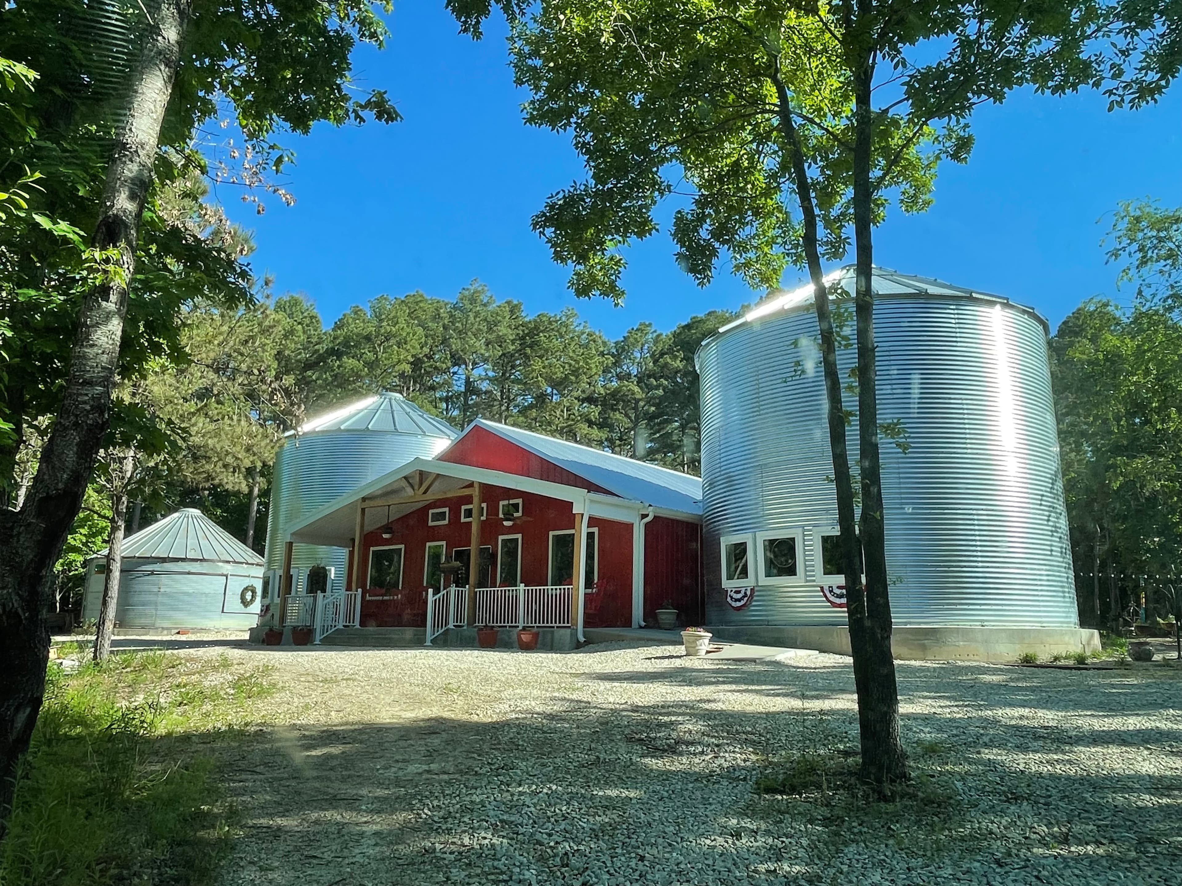 A red barn-style building with a white porch is flanked by two large, silver grain silos that have been converted for use. The property is surrounded by tall trees under a clear blue sky, with gravel covering the ground in front.