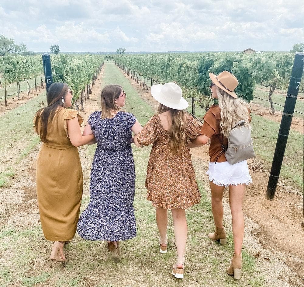 Four women walking hand-in-hand through a vineyard under a cloudy sky.