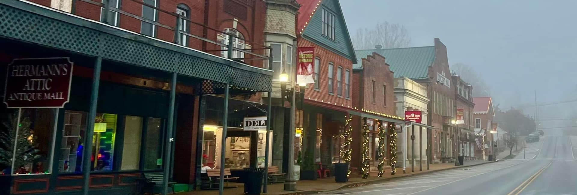 A misty street lined with quaint brick buildings and antique shops, adorned with festive lights.