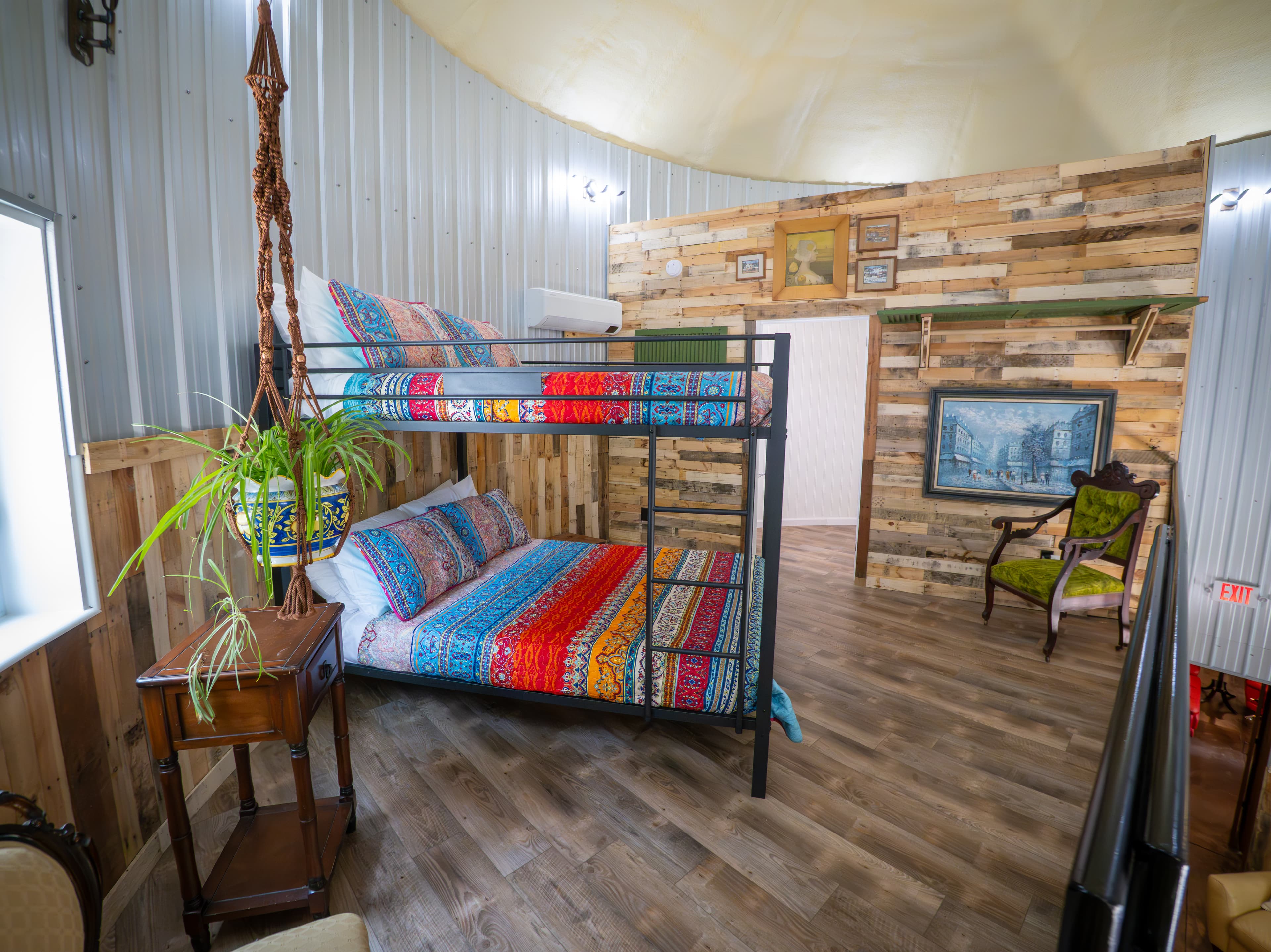 A wide-angle view of an eclectic sleeping loft inside a circular grain bin, featuring a black metal bunk bed with vibrant, colorful patterned linens. The space is detailed with rustic wood-planked and corrugated metal walls, a lime green velvet armchair, and a hanging spider plant in a blue ceramic pot.