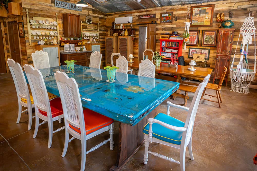 A vibrant, rustic dining area featuring a blue wooden table surrounded by colorful chairs, with wooden decor elements in the background.