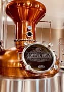 Copper distilling equipment with a branded plaque reading "Copper Muck Distilling."