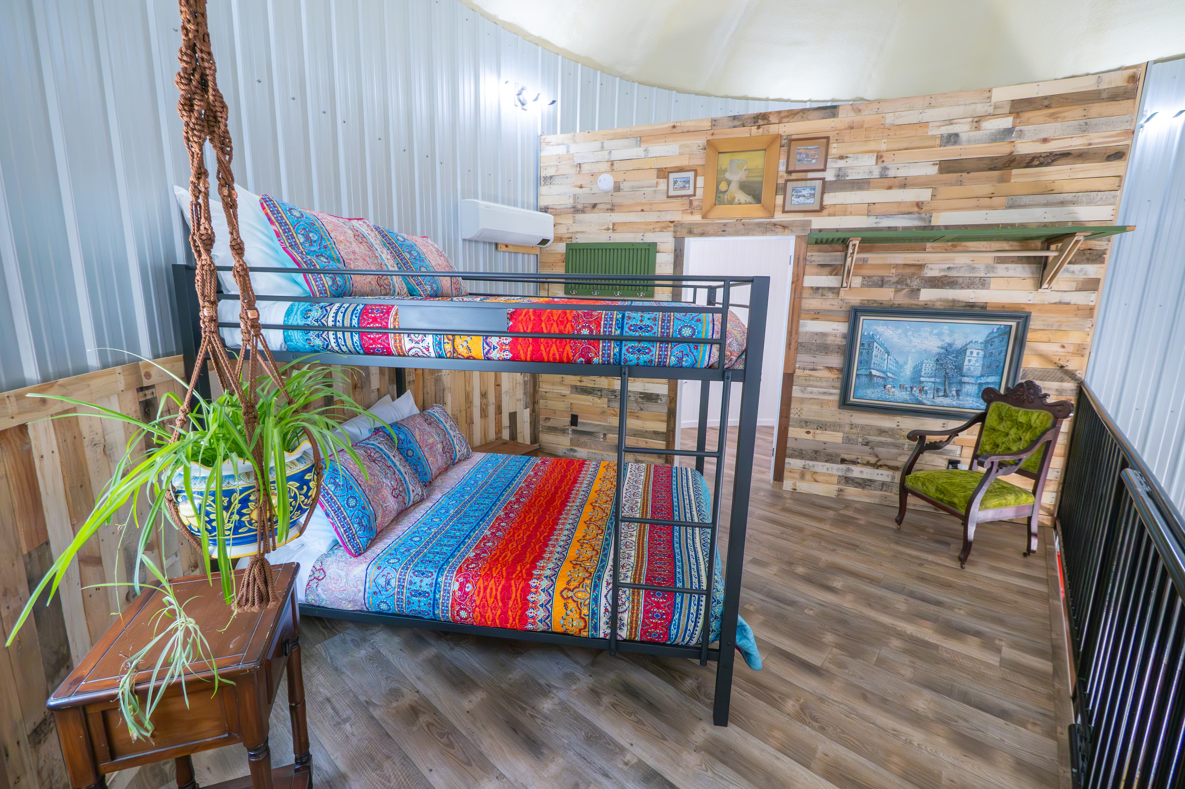 A black metal queen bunk bed with vibrant, multi-colored striped bedding sits on a wood-plank floor in a lofted area. The space features rustic wood-paneled and corrugated metal walls, a hanging plant in a blue patterned pot, and a green velvet armchair in the corner.